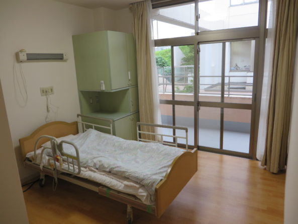 介護施設の個室。ベッド、エアコン、大型窓が配置された明るい居室。